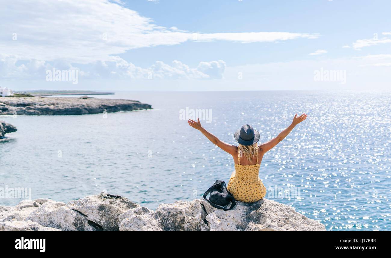 Donna spensierata seduta sulla roccia con le braccia sollevate di fronte al mare in giornata di sole, Minorca, Spagna Foto Stock