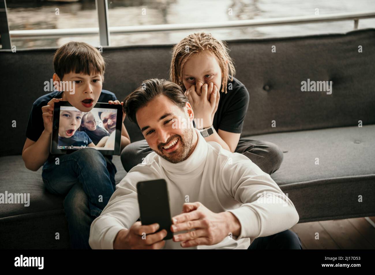 Uomo felice che prende selfie attraverso lo smartphone con figli che fanno volti a casa Foto Stock