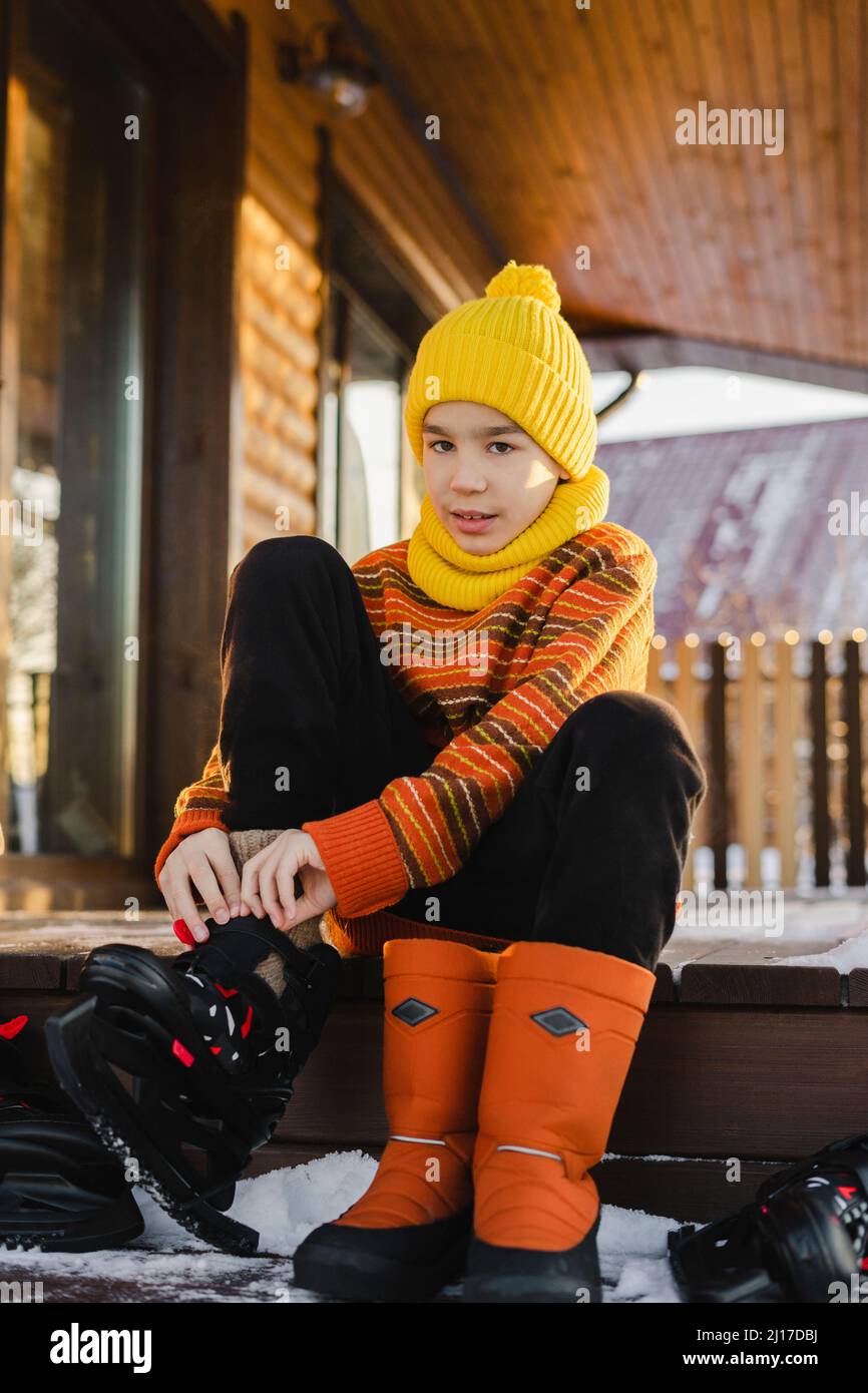 Ragazzo in maglia cappello con pattini da ghiaccio seduti al portico di casa in inverno Foto Stock