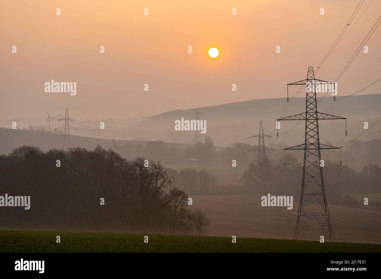 Ledgerwood in Scottish Borders, Regno Unito. 23rd marzo 2022. Energia, energia, elettricità. Il sole sorge in un altro giorno di elevati prezzi dell'energia, mentre il costo della vita continua ad aumentare. La transizione all'energia verde è un tema prioritario, ma anche l'equilibrio tra la riduzione dei costi energetici attualmente elevati a causa della pandemia covida e dell'attuale guerra in Ucraina e le sanzioni contro la Russia. La vista è di tralicci di elettricità e di una turbina eolica a distanza quando il sole sorge vicino a Ledgerwood, ai confini scozzesi. Credit: phil wilkinson/Alamy Live News Foto Stock