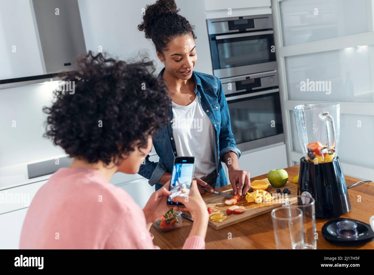 Giovane donna fotografando amico tagliando frutta in cucina a casa Foto Stock