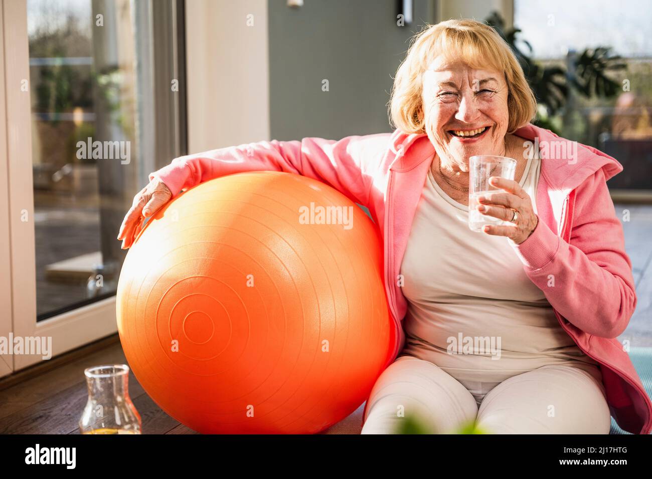 Donna anziana allegra che tiene un bicchiere d'acqua seduta con palla fitness a casa Foto Stock