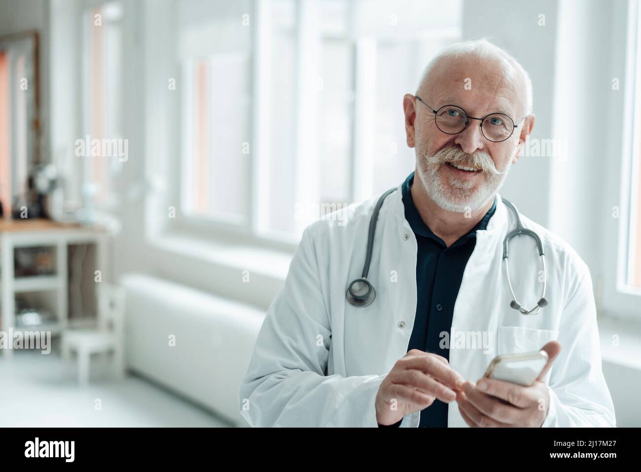 Sorridente medico senior che tiene il telefono cellulare in ospedale Foto Stock