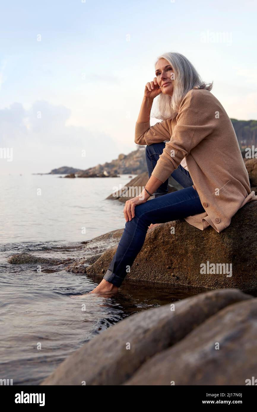 Donna dai capelli grigi seduta su roccia in spiaggia Foto Stock