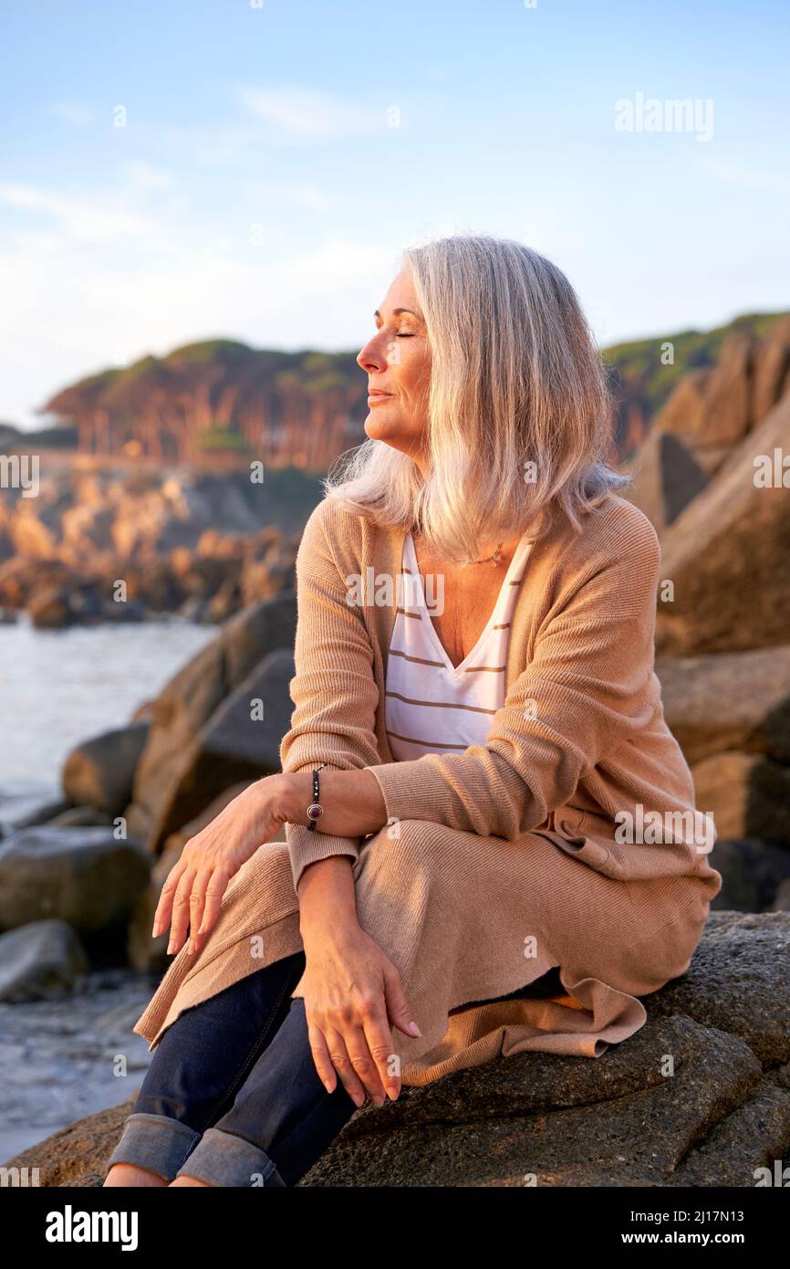 Donna dai capelli grigi con occhi chiusi seduta su roccia in spiaggia Foto Stock