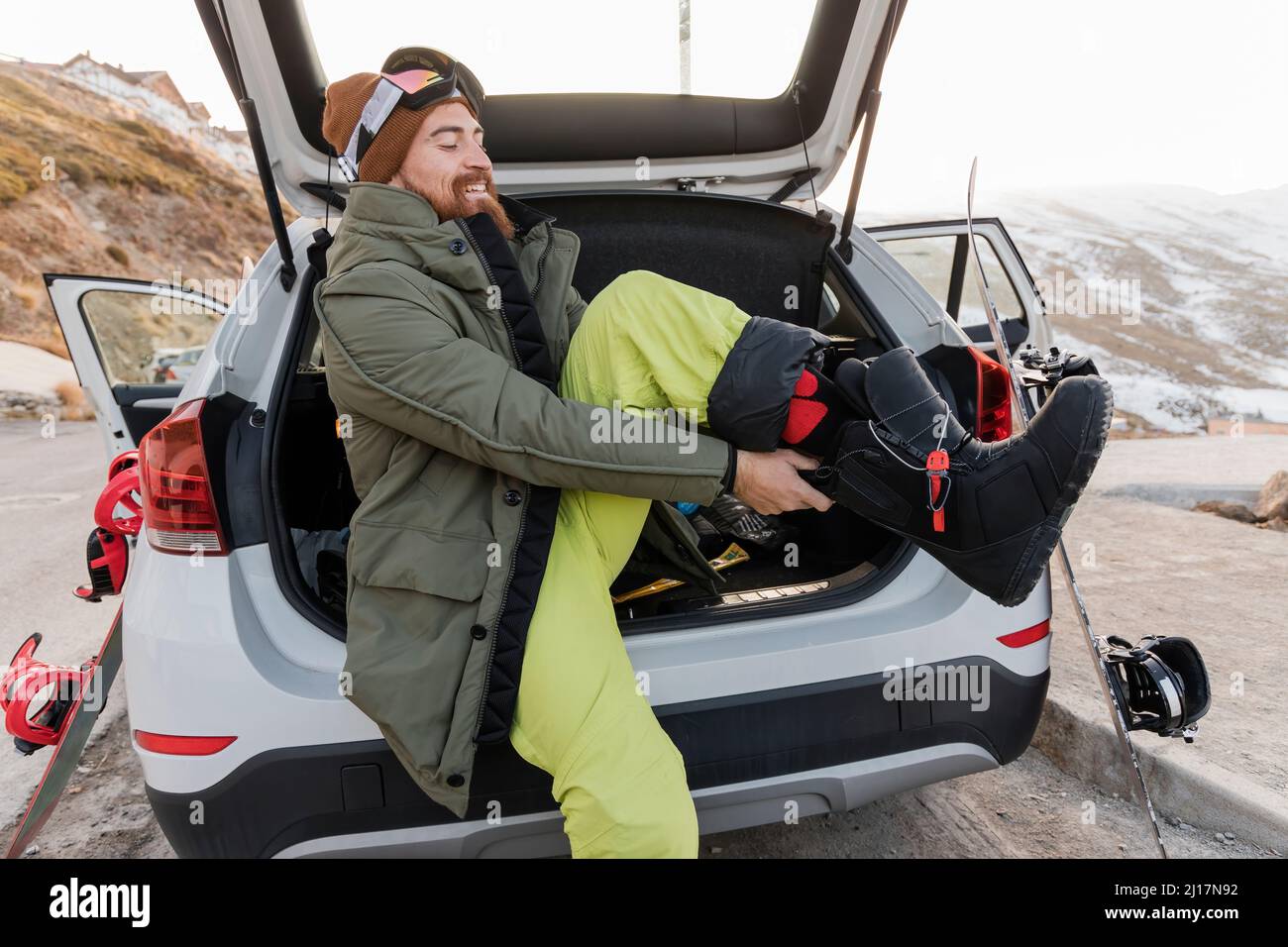 Sorridente giovane uomo nel bagagliaio dell'auto che indossa gli scarponi da sci Foto Stock