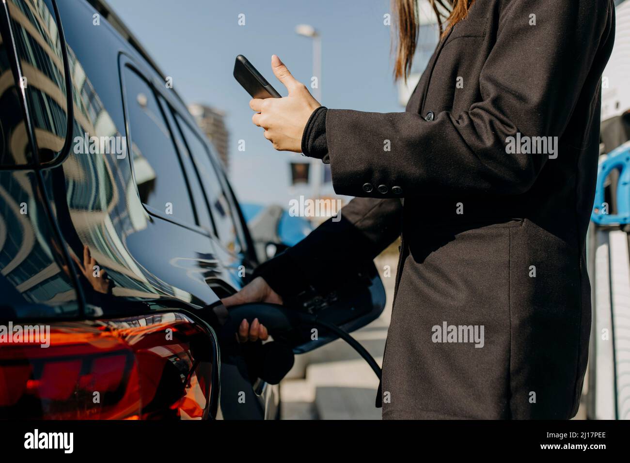 Donna che utilizza uno smartphone vicino all'auto mentre viene caricata al punto di ricarica Foto Stock