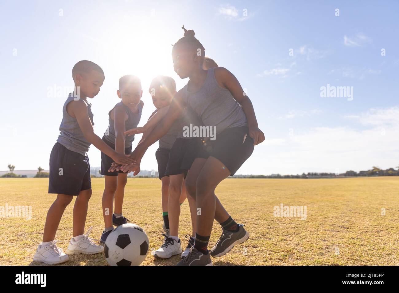 Ragazzi di scuola elementare multirazziale accatastano mani su pallone da calcio su campo contro cielo in giornata di sole Foto Stock