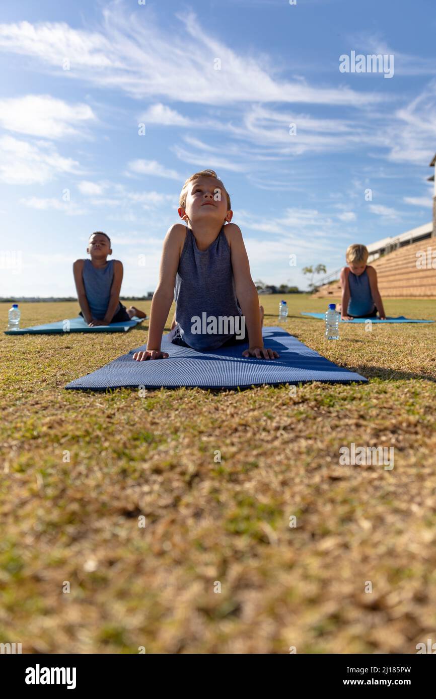 I ragazzi della scuola elementare multirazziale che fanno il cane rivolto verso l'alto pongono l'esercizio sul tappeto di yoga in terra Foto Stock