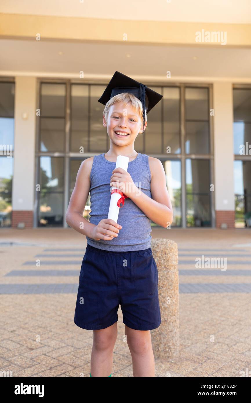 Sorridente scolaretto caucasico elementare con mortarboard e laurea in piedi contro l'edificio scolastico Foto Stock