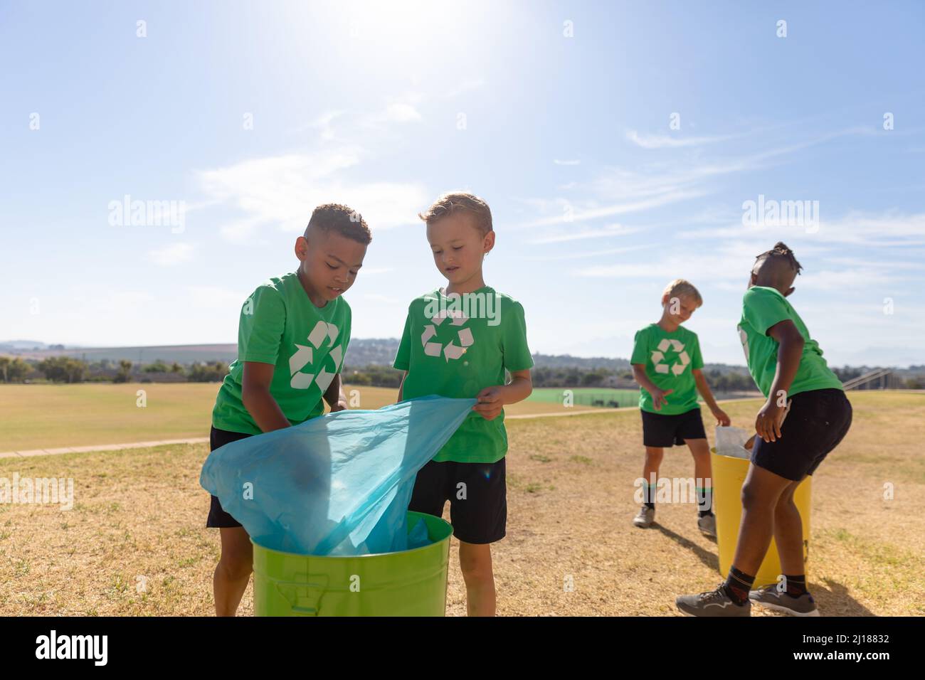 Ragazzi di scuola elementare multirazziale che mettono sacchi di rifiuti in cestino sulla terra della scuola contro il cielo Foto Stock