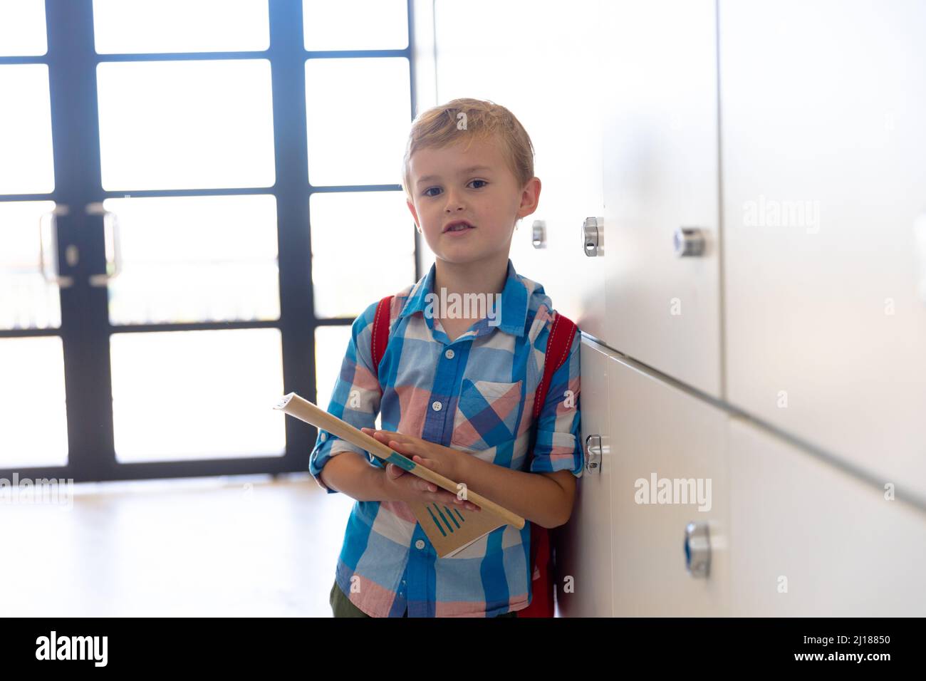 Ritratto di carino scolaro caucasico elementare con libro in piedi da armadietti a scuola Foto Stock