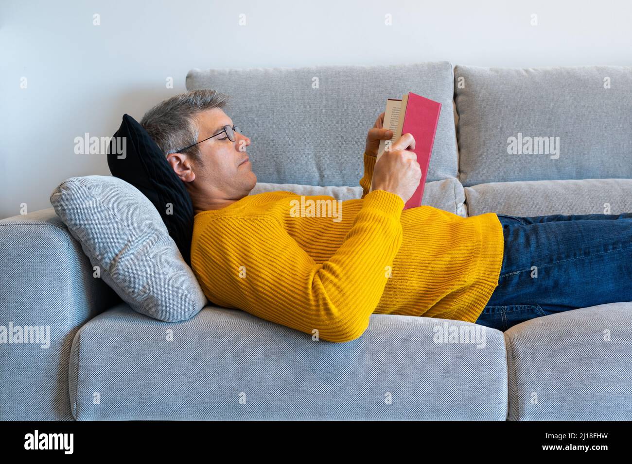Uomo rilassato di mezza età con occhiali sdraiati sul divano leggendo un libro. Comfort a casa tua Foto Stock