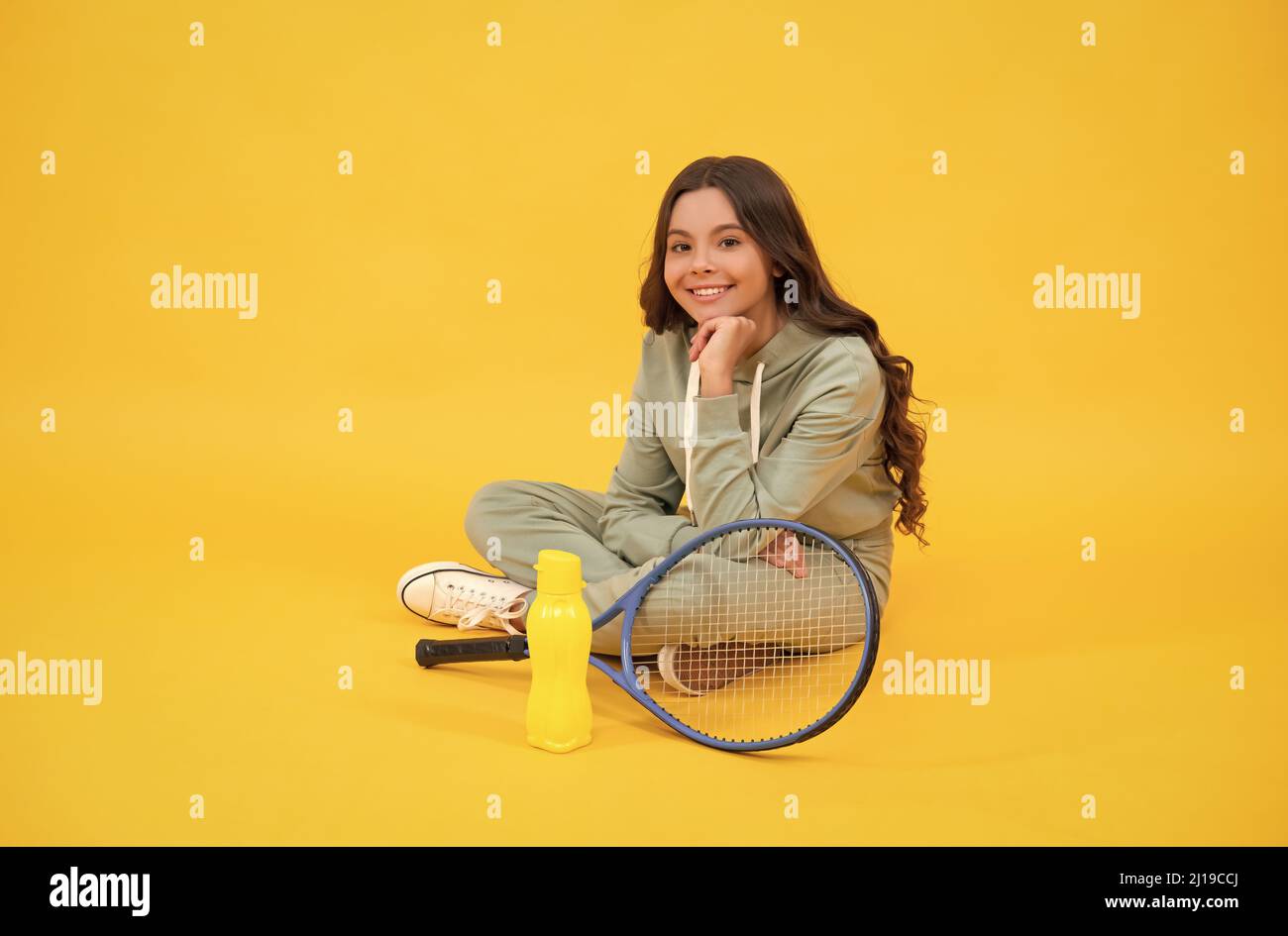 bambino allegro seduto in abbigliamento sportivo con racchetta di badminton e bottiglia d'acqua su sfondo giallo, relax Foto Stock