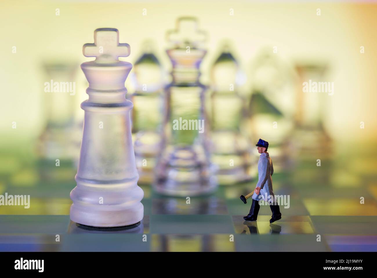 Scacchiera di vetro e pezzi. Uomo in miniatura con martello che cammina verso re sfondo caldo Foto Stock