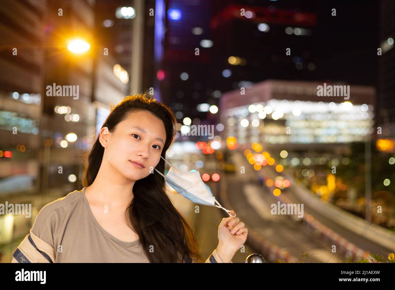 Bella giovane studentessa università rimuovere la maschera per respirare aria fresca nel centro, Hong Kong con vista notturna di sfocato luci città colorate in bac Foto Stock