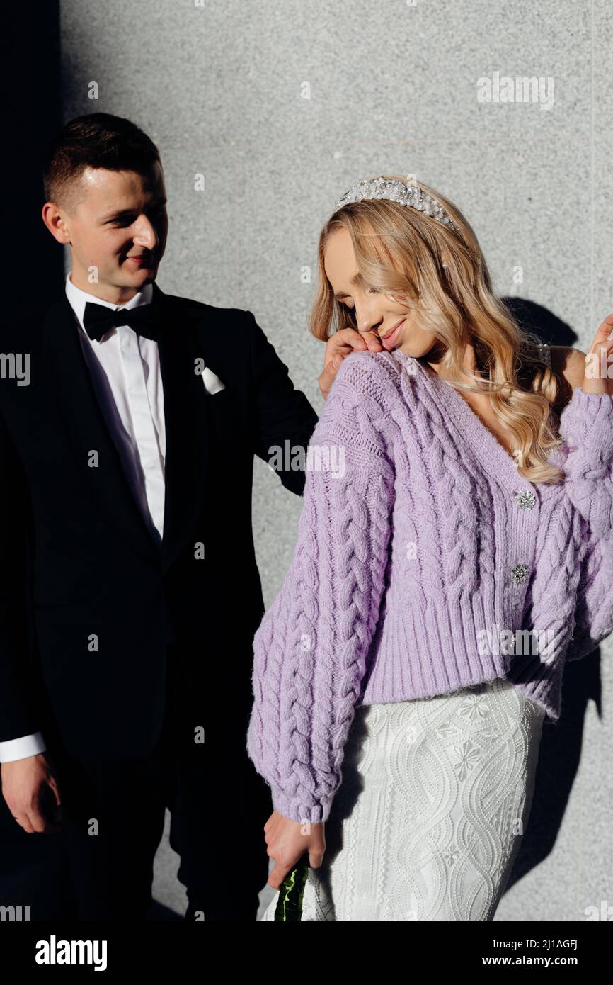 Giovane sposa perfetta in abito da sposa, cardigan lilla e bandeau in testa in piedi con schiena a sposo in abito nero. Foto Stock