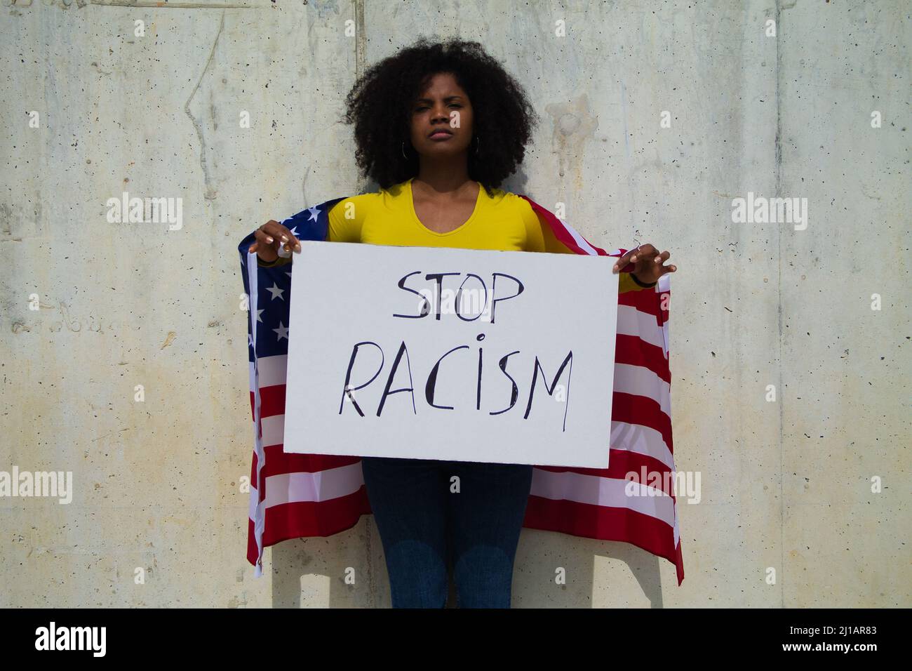 Una donna afroamericana tiene in mano una bandiera contro il razzismo e la bandiera degli stati uniti sulle spalle. In sfondo grigio parete Foto Stock