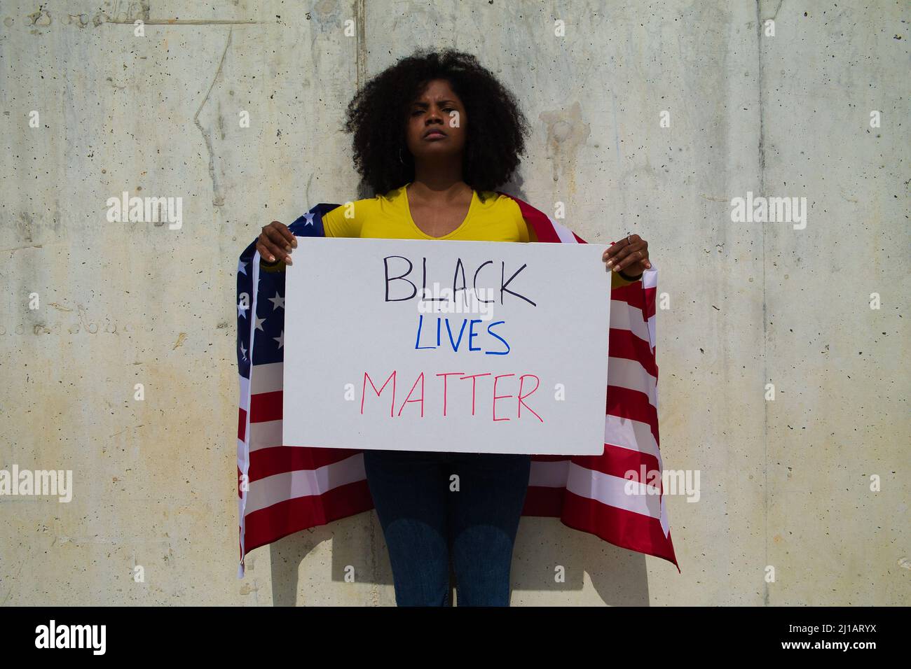 Una donna afroamericana tiene una materia nera della vita nelle sue mani e la bandiera degli stati uniti sopra la sua spalla. In sfondo grigio parete Foto Stock