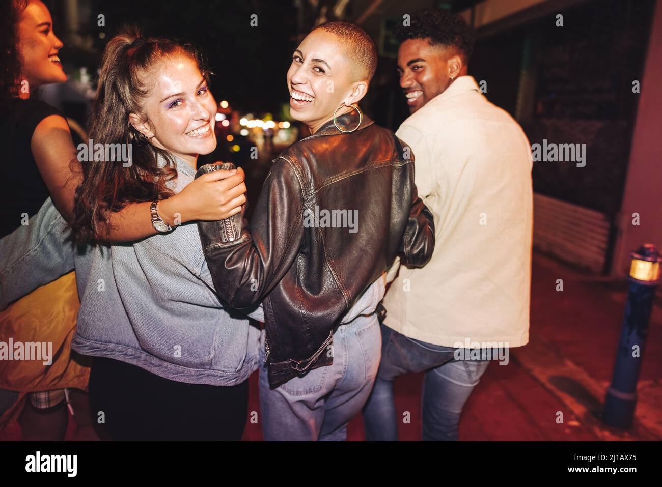 Buon tempo il sabato sera. Vista posteriore di un gruppo di amici vibranti ridere felicemente mentre camminano in città di notte. Patatine fritte multiculturali Foto Stock