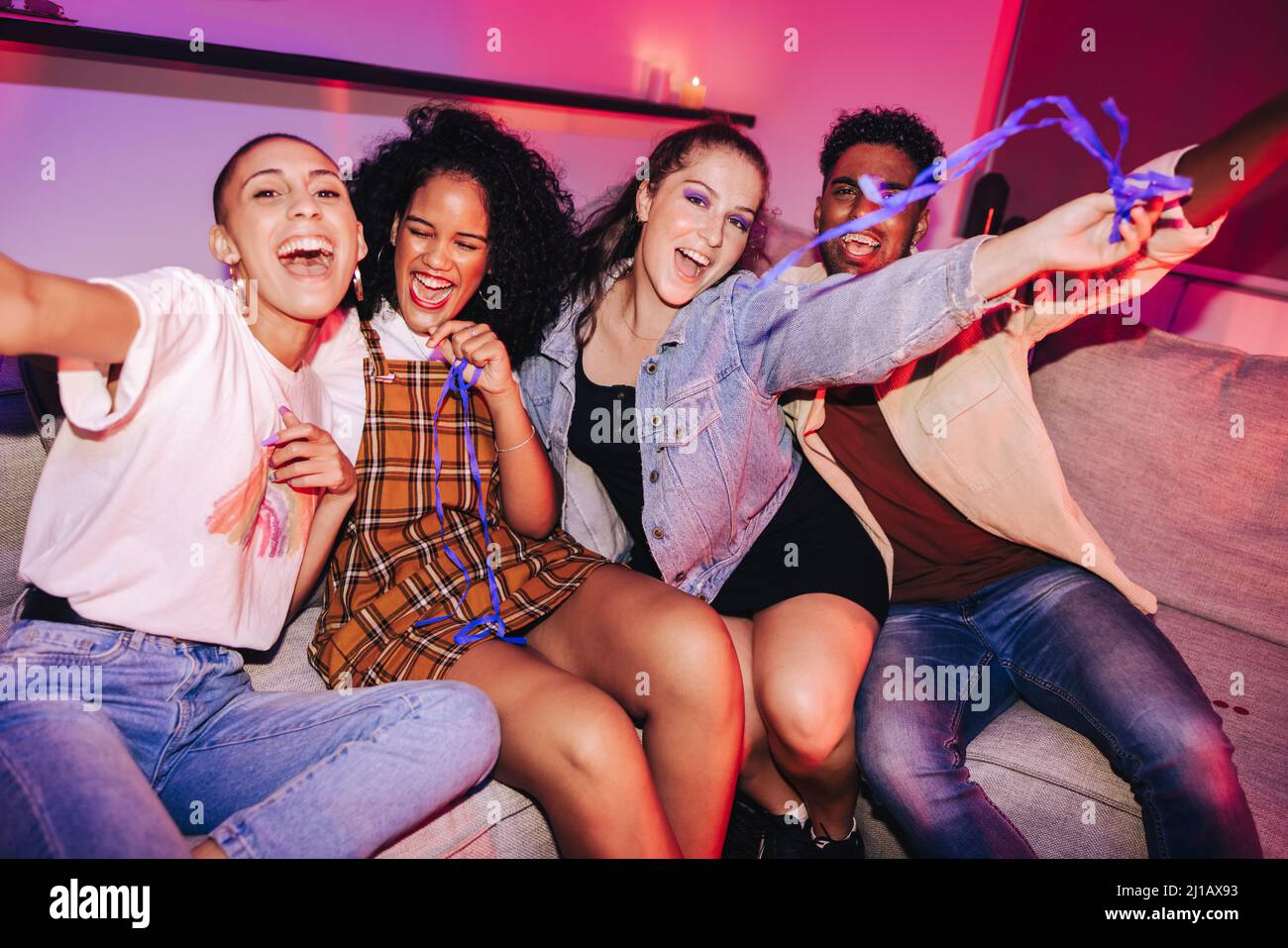 Gruppo di amici che festeggiano in una festa della casa. I giovani vibranti si divertono seduti su un divano in una luminosa luce al neon. Amici multiculturali en Foto Stock