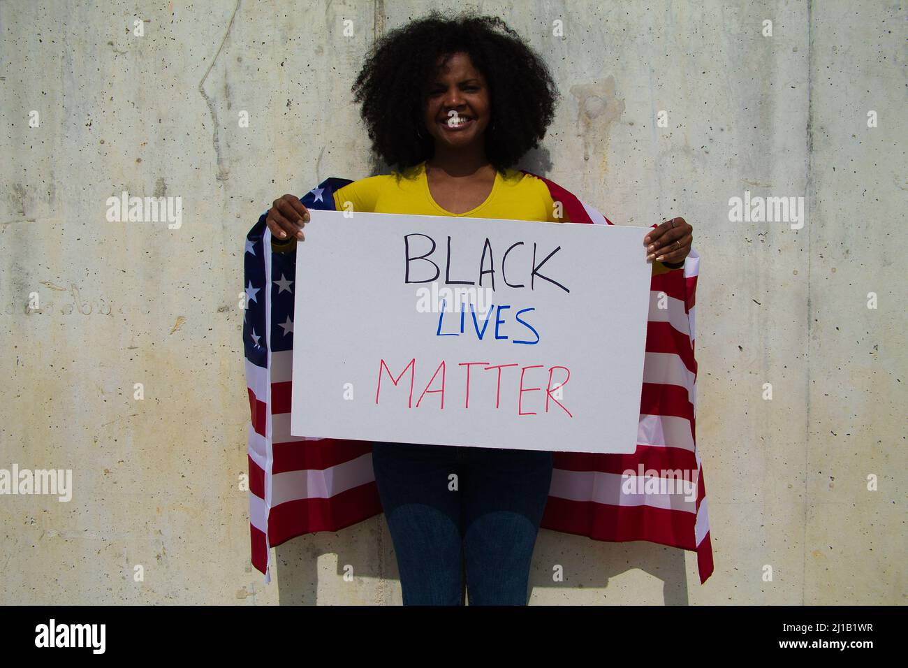 Una donna afroamericana tiene una materia nera della vita nelle sue mani e la bandiera degli stati uniti sopra la sua spalla. In sfondo grigio parete. Foto Stock