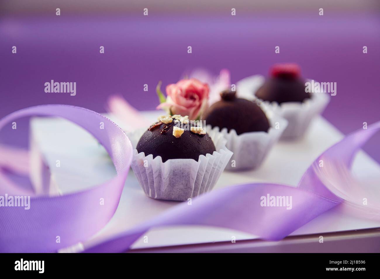 Delicate caramelle al cioccolato festive. Piume rosa, rosa rosa e nastro viola. Sfondo con motivo di colore viola. Foto Stock