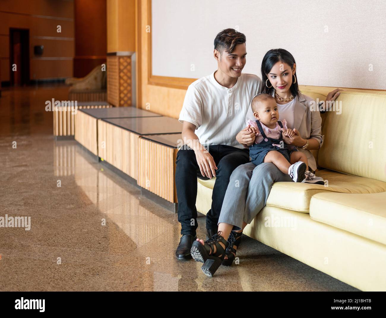 Malay Malese musulmano giovane coppia con una figlia di un anno seduta su una lunga sedia marrone chiaro Foto Stock