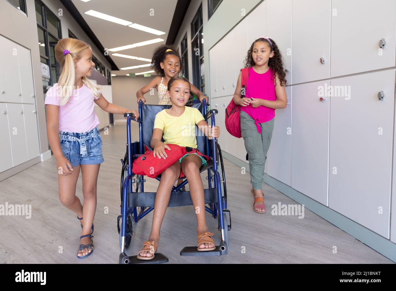 Sorridente studentesse multirazziali che camminano con la compagna di classe femminile seduta su sedia a rotelle Foto Stock