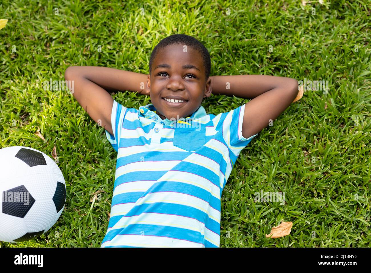 Sorridente ragazzo afroamericano sdraiato con le mani dietro la testa da palla di calcio su erba nel cortile Foto Stock