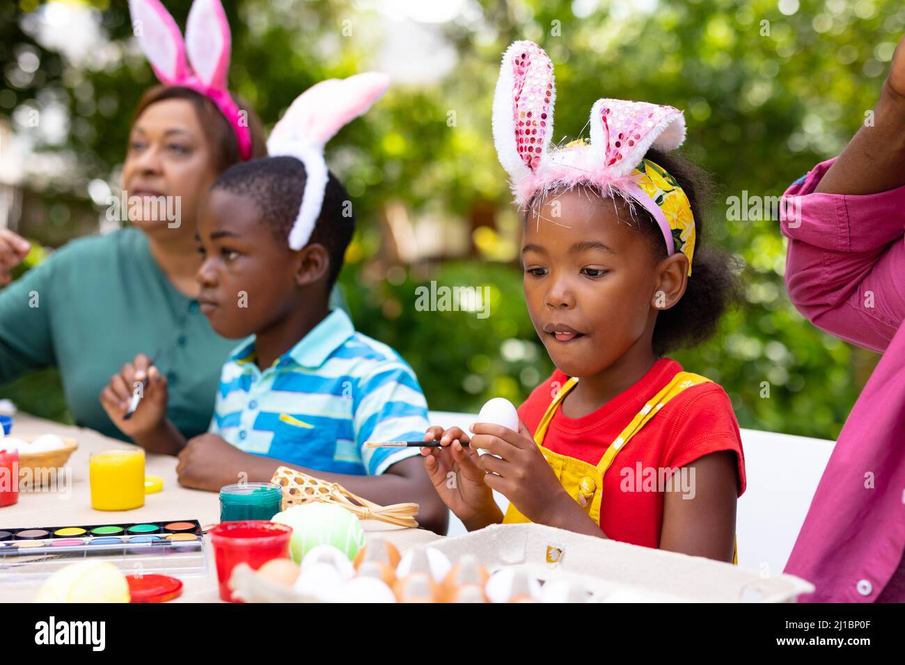 Ragazza afroamericana che indossa le orecchie di coniglio dipingendo le uova con la famiglia in cortile il giorno di pasqua Foto Stock