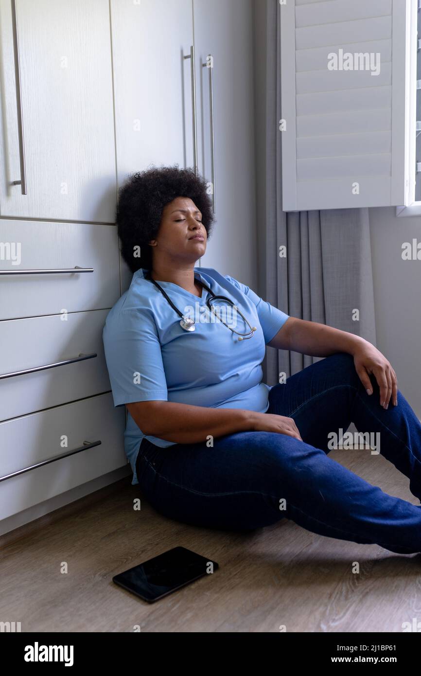 Stressante infermiera femminile afroamericana per adulti di media età con occhi chiusi seduti sul pavimento in ospedale Foto Stock