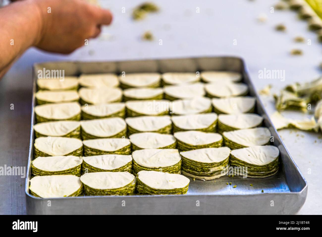 Un vassoio di metallo contenente molte file di pezzi di pasticceria a strati a forma di ventaglio ricoperti di pistacchi si trova nella manifattura di Patisserie Petek Pastahanesi a Gazimağusa/Famagusta, Repubblica Turca di Cipro del Nord (RTNC); una mano entra nel quadro dall’alto, e scarti di impasto sparsi giacciono sulla superficie di lavoro a destra. La pasticceria è stata fondata nel 1976 da Mehmet Bey e Seher Hanım, è considerata oggi una delle più antiche panetterie di Cipro e impiega circa 130 persone. Foto Stock