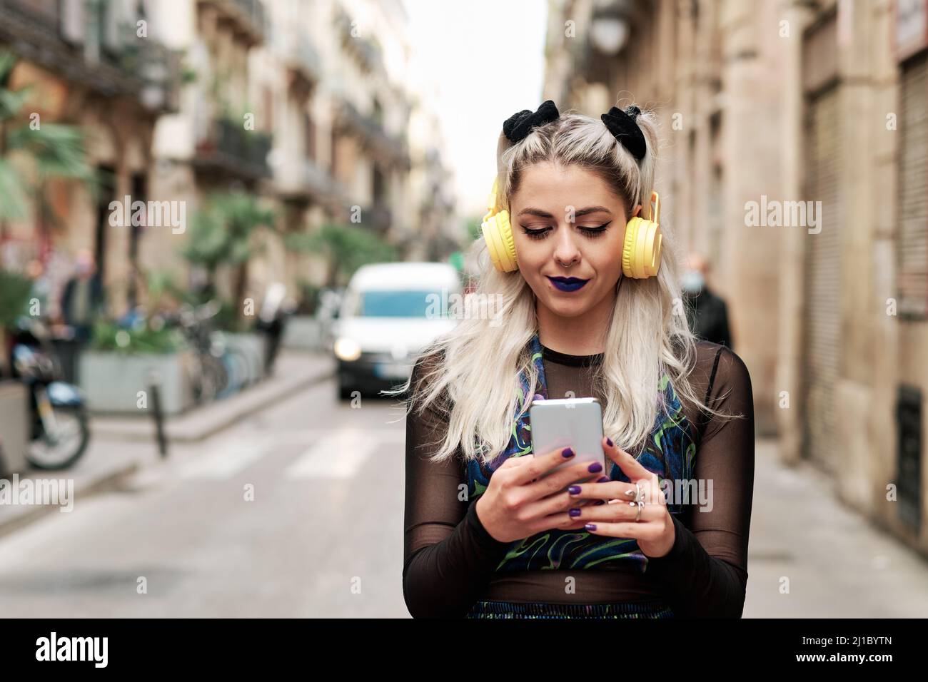 Giovane donna alternativa che usa un telefono cellulare con cuffie mentre cammina lungo la strada. Foto Stock