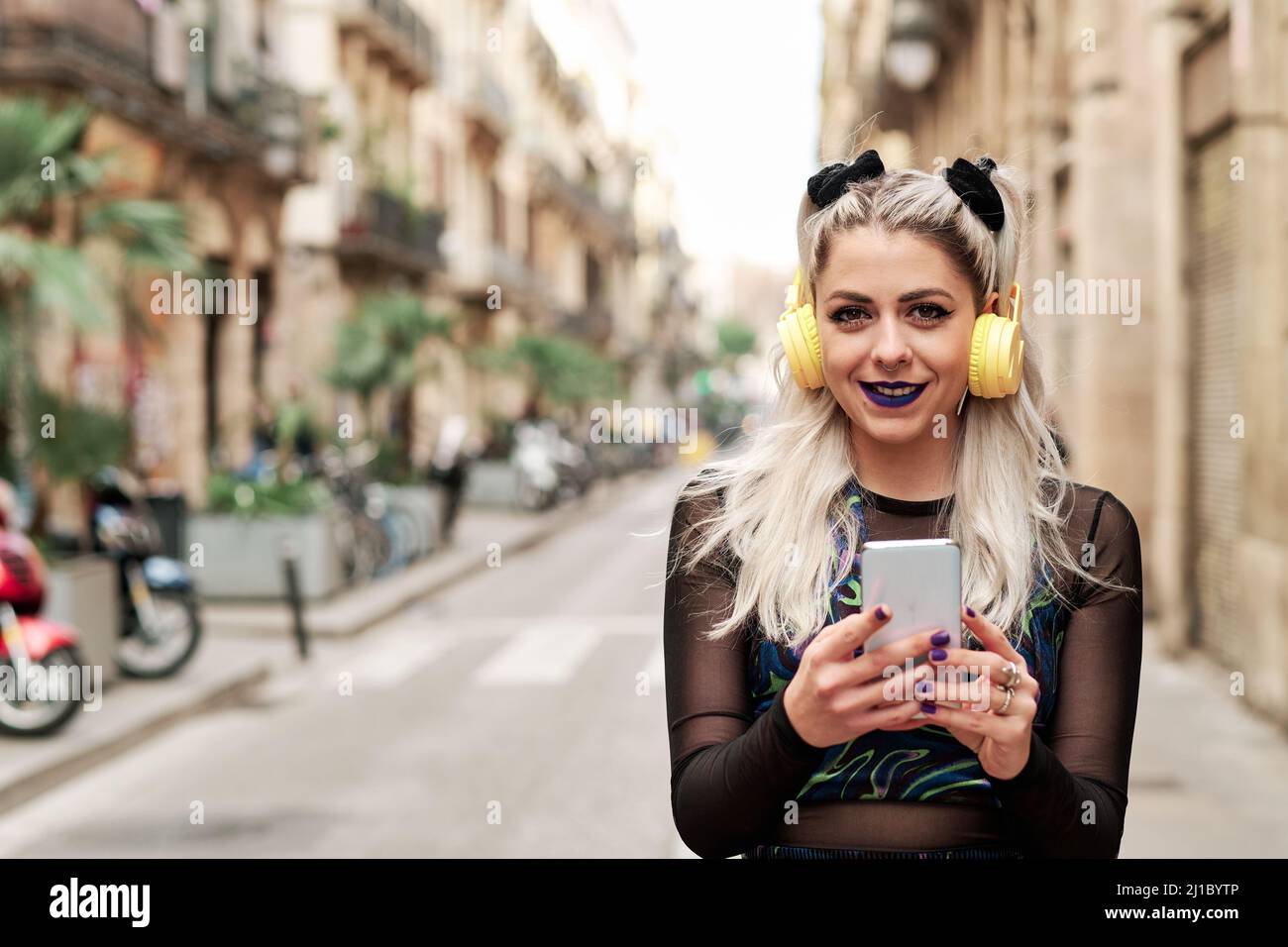 Donna alternativa sorridente mentre ascolti la musica con un telefono cellulare e le cuffie per strada. Foto Stock