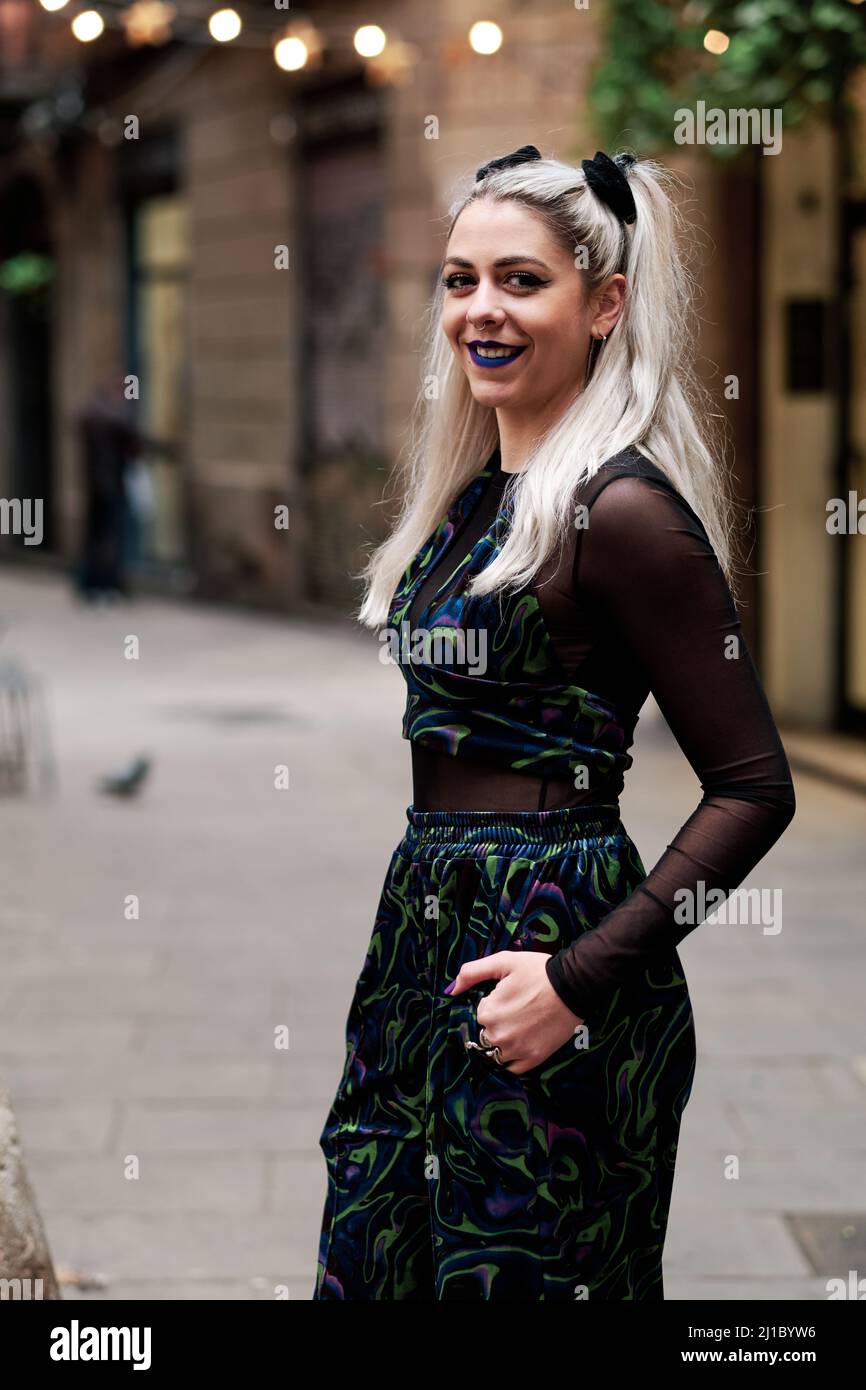 Giovane donna alternativa che guarda la macchina fotografica e sorride mentre si posa all'aperto per strada. Foto Stock