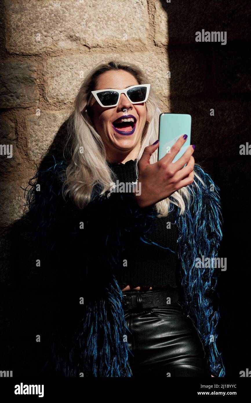 Elegante donna alternativa in occhiali da sole ridendo mentre si fa videochiamata sul cellulare all'aperto. Foto Stock