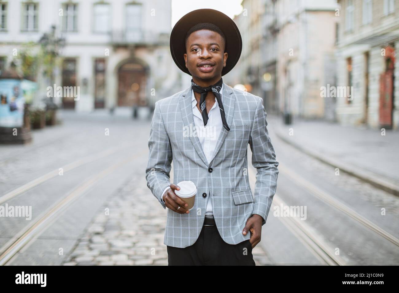 Uomo afroamericano sorridente in piedi sulla strada della città e tenere  una tazza di caffè-to-go. Giovane ippica maschile con cappello nero, abito  elegante e sciarpa sul collo Foto stock - Alamy