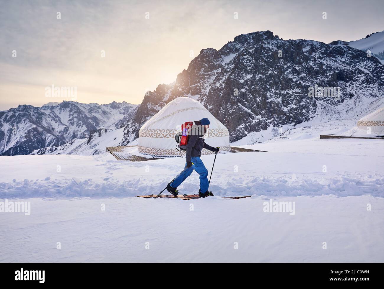 Sciatore uomo sci alpinismo in salita con barba e zaino in alta montagna innevata vicino alla casa tradizionale nomad yurt presso la stazione sciistica di Shymbulak in Almaty, Kazak Foto Stock
