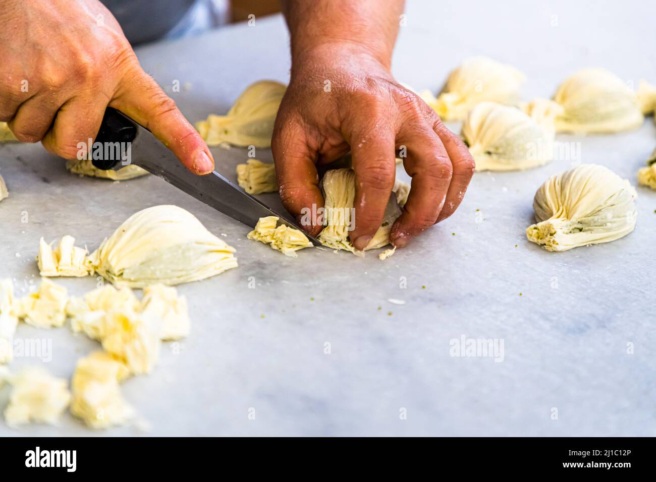 Due mani tagliano l'eccesso di impasto con un coltellino da pezzi di pasta appena modellati a forma di cozza – forse la preparazione del Midye baklava – nella manifattura di Patisserie Petek Pastahanesi a Gazimağusa/Famagusta, Repubblica Turca di Cipro del Nord (RTNC). La pasticceria è stata fondata nel 1976 da Mehmet Bey e Seher Hanım, è oggi considerata una delle più antiche panetterie di Cipro e impiega circa 130 persone. Foto Stock