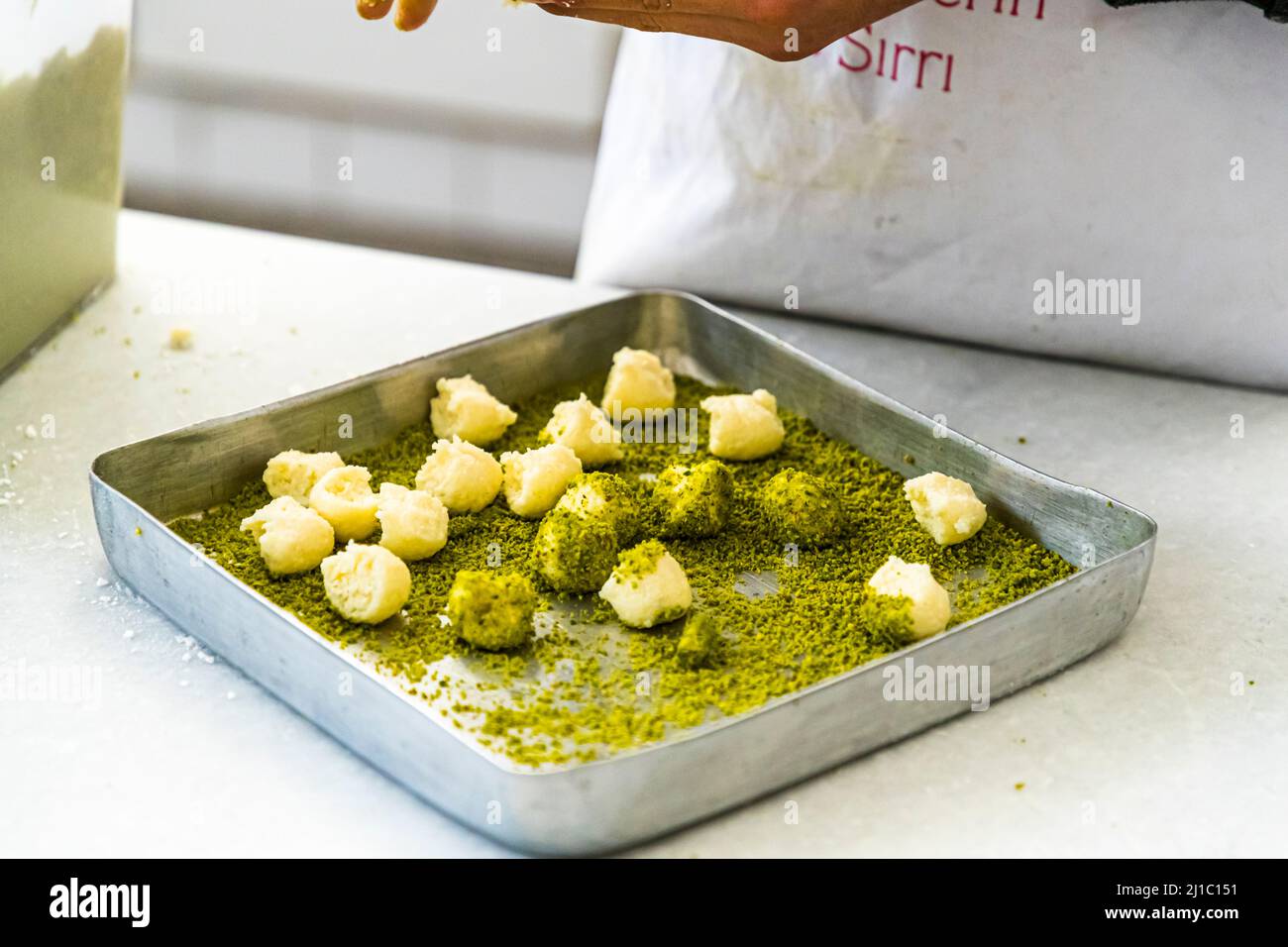 Piccole palline rotonde di impasto riempiono una teglia quadrata, disposte vicine, alcune già arrotolate nella pasta di pistacchi, mentre una mano sparge ulteriori pistacchi dall'alto, nella manifattura della Patisserie Petek Pastahanesi a Gazimağusa/Famagusta, Repubblica Turca di Cipro del Nord (RTNC). La pasticceria è stata fondata nel 1976 da Mehmet Bey e Seher Hanım, è considerata oggi una delle più antiche panetterie di Cipro e impiega circa 130 persone. Foto Stock