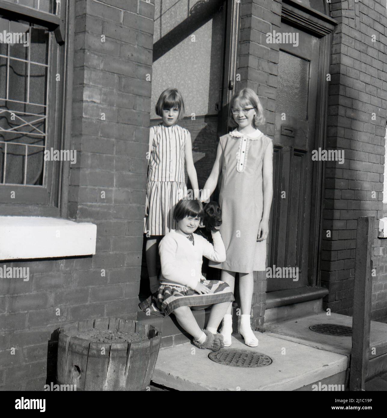 1950s, storica, tre ragazze giovani con il cane di famiglia in piedi alla porta della loro casa terrraced vittoriana in una strada acciottolata a Stockport, Manchester, Inghilterra, Regno Unito. Notare il coperchio in ferro per il foro di carbone nel gradino anteriore Foto Stock