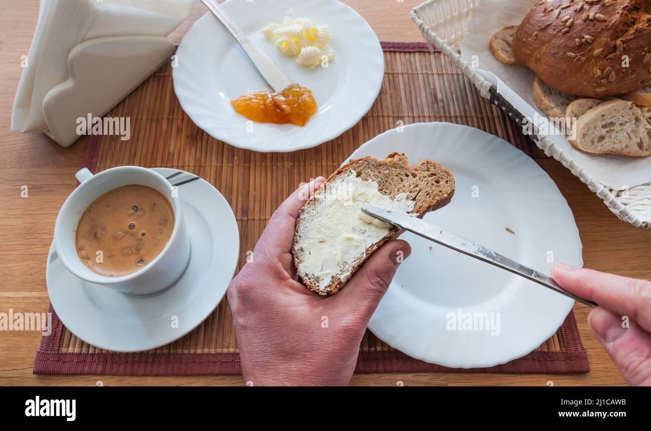 Colazione italiana con pane, burro e marmellata. Le mani dell'uomo in primo piano stendono il burro su una fetta di pane con un coltello Foto Stock