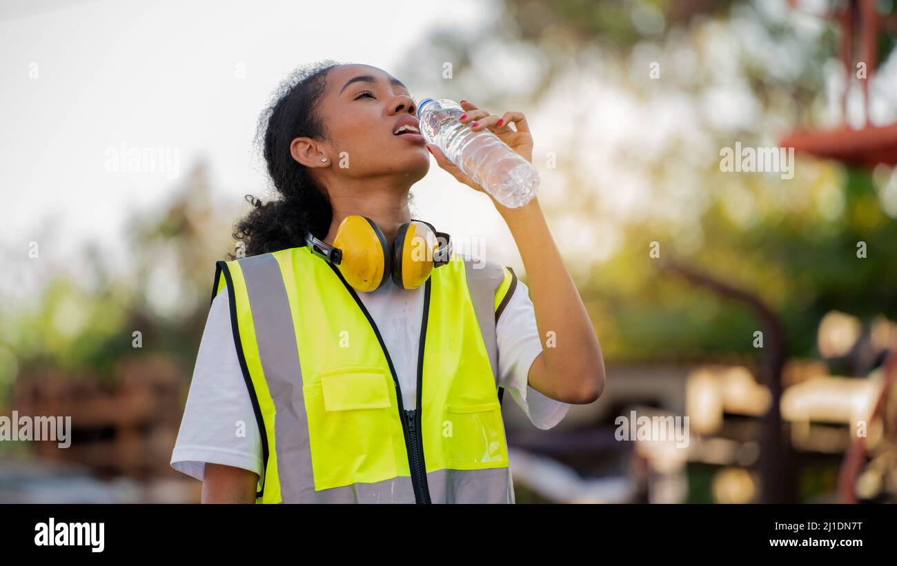 Divergente Una donna afroamericana uomo o lavoratore che indossa la sicurezza degli occhi incrociati sta bevendo una bottiglia d'acqua dopo il lavoro di completamento e rilassarsi sulla o. Foto Stock