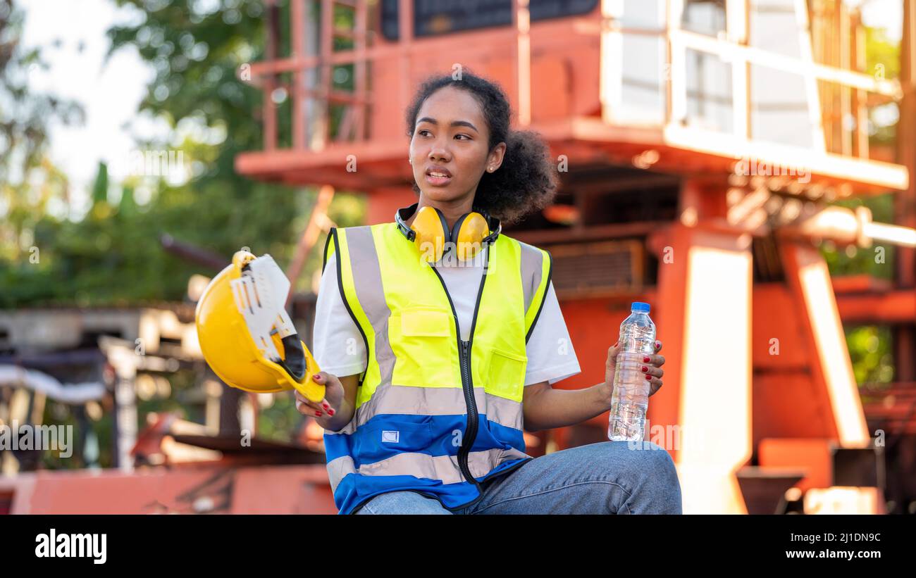 Divergente Una donna afroamericana uomo o lavoratore che indossa la sicurezza degli occhi incrociati sta bevendo una bottiglia d'acqua dopo il lavoro di completamento e rilassarsi sulla o. Foto Stock