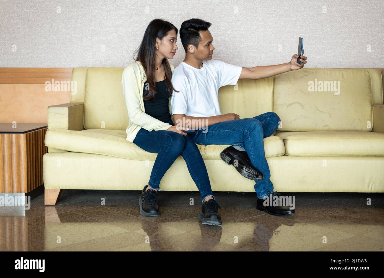 Malay Asian Malese musulmana coppia felice prendere una foto selfie utilizzando un telefono a mano, mentre seduto su una sedia marrone chiaro divano divano divano a casa Foto Stock