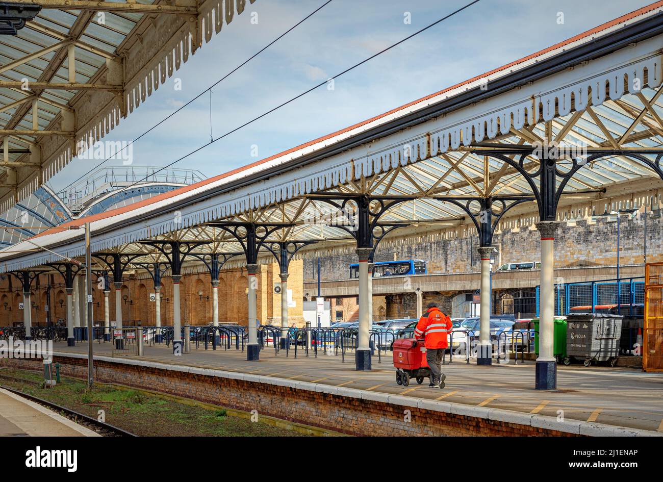 Un postino spinge un carrello lungo una piattaforma della stazione ferroviaria. Uno storico baldacchino del 19th secolo è in testa e una strada con traffico è sullo sfondo. Foto Stock
