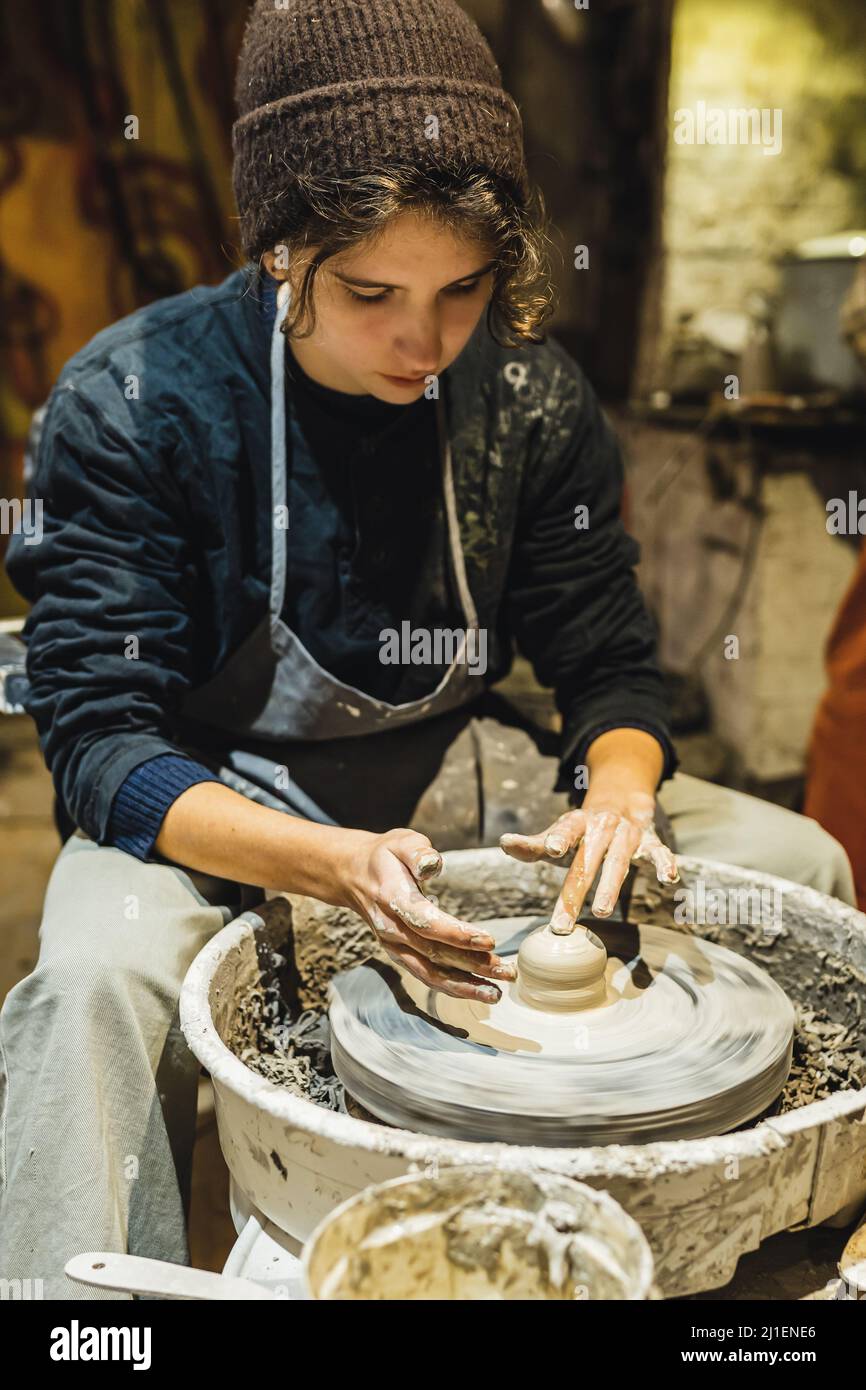 adolescente in abiti da lavoro macchiati con argilla sta studiando ceramica, imparando a lavorare alla ruota di vasaio nel laboratorio di vasaio. Foto Stock