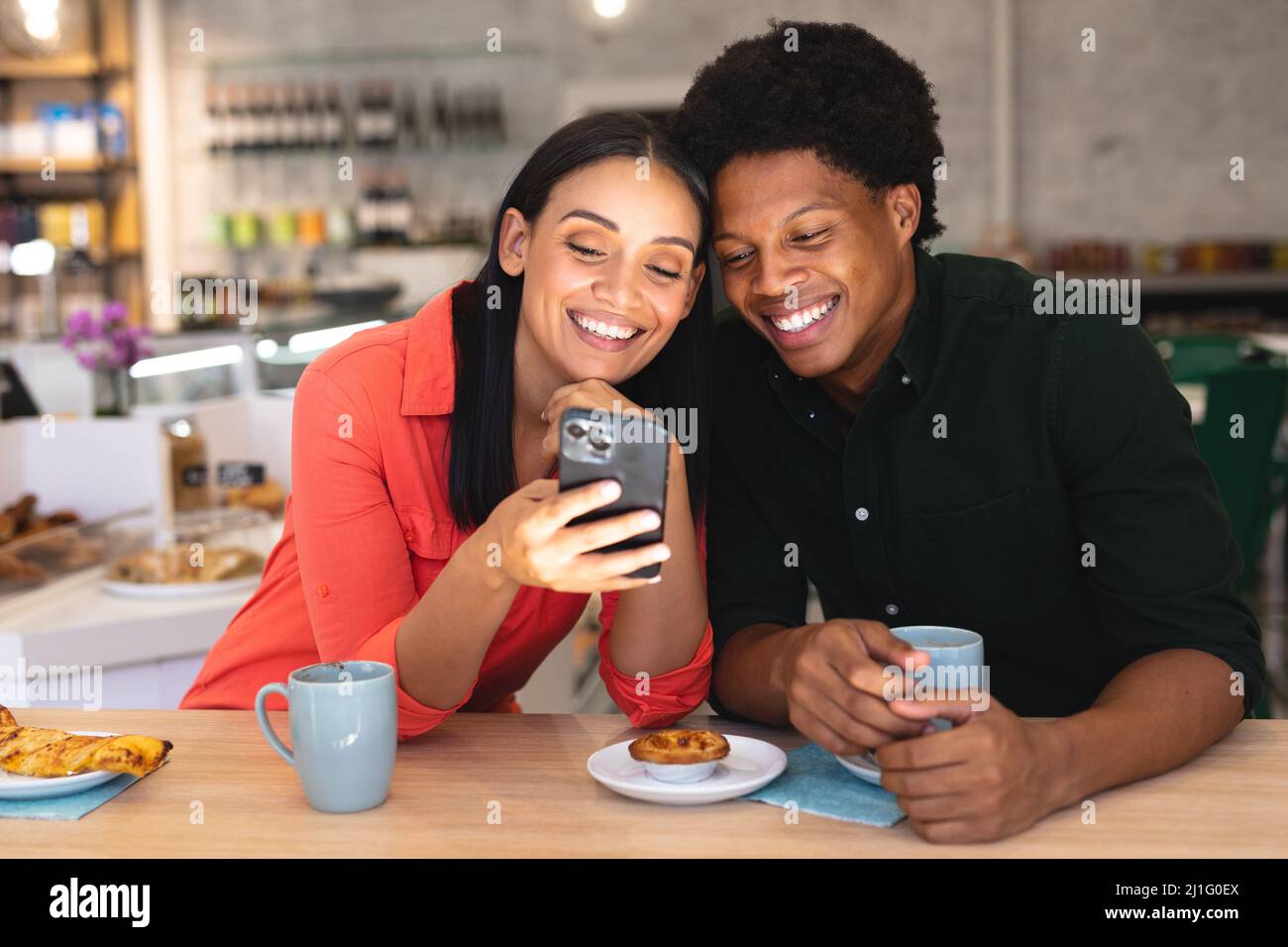 Felice giovane coppia multirazziale condividere smartphone mentre si siede con caffè insieme in caffè. Inalterato, caffè cultura, persone, amore, tecnologia e li Foto Stock