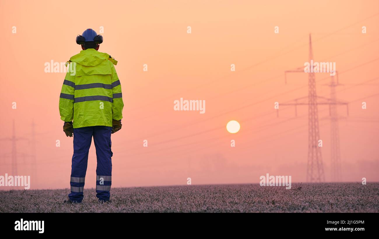 Vista posteriore dell'ingegnere elettrico contro i tralicci di elettricità durante la gelida mattina d'inverno. Temi dell'approvvigionamento energetico, dell'industria e dell'ambiente. Foto Stock